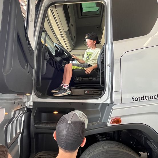 Ein junger Junge sitzt auf dem Fahrersitz eines weißen Ford-Lkws, mit offener Tür. Eine andere Person, die eine Kappe trägt, beobachtet von unten.