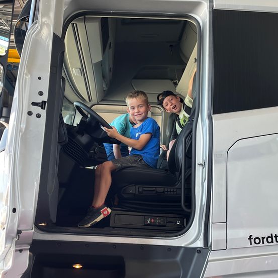 Zwei junge Jungen sitzen auf dem Fahrer- und Beifahrersitz eines weißen Ford-Lkws mit offener Tür. Ein Junge trägt ein blaues T-Shirt und Shorts, der andere ein schwarzes T-Shirt und eine Mütze.