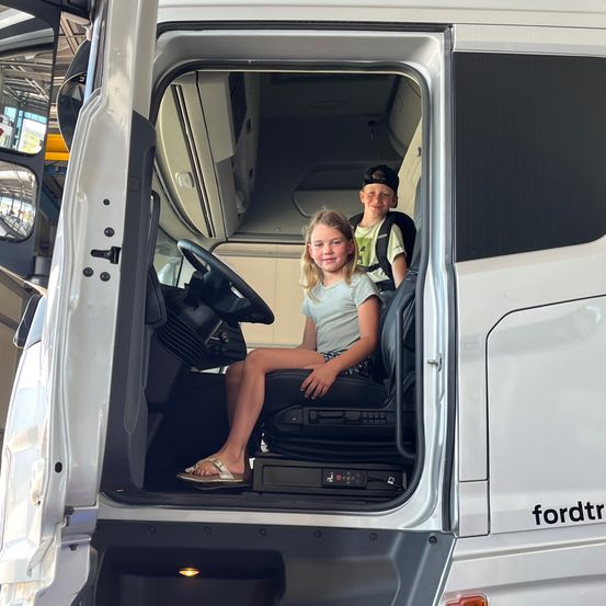 Zwei Kinder sitzen in einem weißen Fahrzeug, ein Mädchen in Sandalen und ein Junge mit Rucksack. Die Fahrzeugtür ist geöffnet und zeigt den Innenraum.