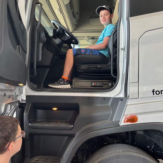 Ein junger Junge sitzt auf dem Fahrersitz eines weißen Lastwagens. Ein anderer Junge mit Brille schaut von unten zu ihm auf.