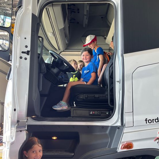 Im Inneren eines Ford-Lkws sitzen mehrere Kinder auf dem Fahrersitz, tragen Mützen und Turnschuhe. Ein Mädchen lächelt und schaut nach vorne, während ein Junge neben ihr etwas zu betrachten scheint. Ein weiterer Junge ist hinter ihnen und schaut möglicherweise aus dem Fenster.