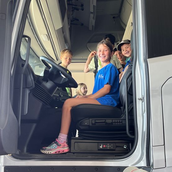 Fünf Kinder sitzen in einem Bus, ein Mädchen lächelt, andere schauen aus dem Fenster. Der Fahrersitz ist mit dem Lenkrad sichtbar.