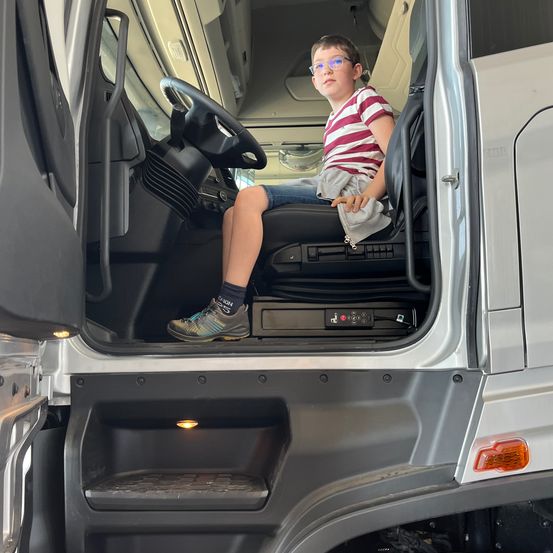 Ein Junge mit Brille und gestreiftem Shirt sitzt im Fahrersitz eines Lastwagens und hält ein Handtuch.