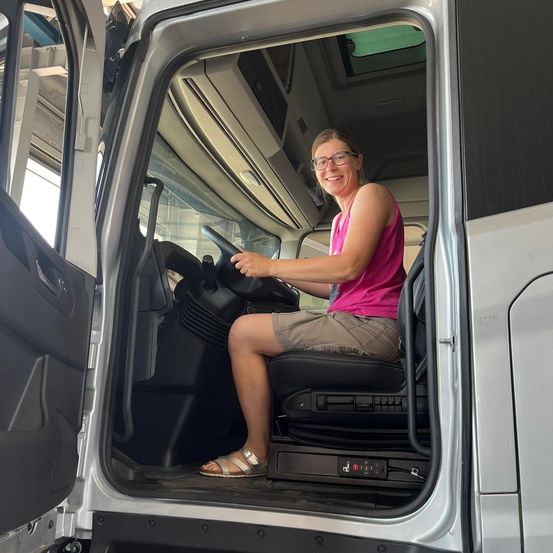 Eine Frau sitzt im Fahrersitz eines Busses, trägt eine Brille und ein rosa Oberteil und lächelt für ein Foto. Der Bus hat offene Türen und das Innere ist sichtbar.