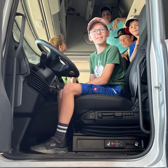 Ein Junge in einem grünen T-Shirt sitzt auf dem Fahrersitz eines Vans, während mehrere andere Kinder hinter ihm sitzen. Die Tür des Vans ist geöffnet, und der Fahrersitz ist sichtbar.