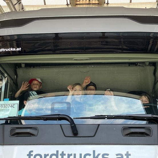 Im Inneren eines Ford-Lkws sitzen mehrere Kinder. Sie scheinen begeistert zu winken. Der Lkw hat den Text 'fordtrucks.at' auf der Vorderseite.