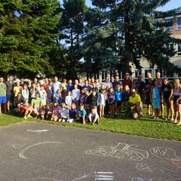 Eine große Gruppe von Menschen, darunter Kinder, versammelt sich auf einem Rasen und posiert lächelnd für ein Foto. Sie stehen vor einem Gebäude mit Bäumen und einer Kreidezeichnung auf dem Boden.