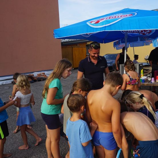 Eine Gruppe von Kindern und einem Mann versammelt sich unter einem blauen Schirm. Sie sind draußen in der Nähe eines Tisches und eines Gebäudes. Der Mann steht und die Kinder schauen sich etwas an.
