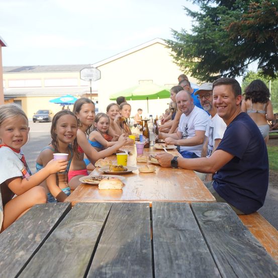 Eine Gruppe von Menschen, darunter Erwachsene und Kinder, sitzen um einen Holzpicknicktisch herum. Einige essen und es gibt Teller mit Essen und Getränken. Hinter ihnen ist ein Schirm und ein Gebäude zu sehen.