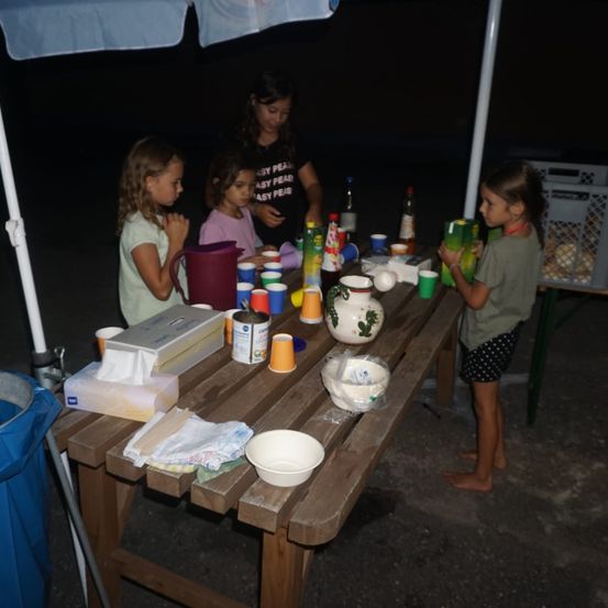Vier Mädchen stehen bei Nacht unter einem Regenschirm an einem Tisch. Der Tisch ist mit Tassen, Schüsseln, Flaschen und anderen Gegenständen gefüllt. Ein Mädchen schaut etwas auf dem Tisch an.