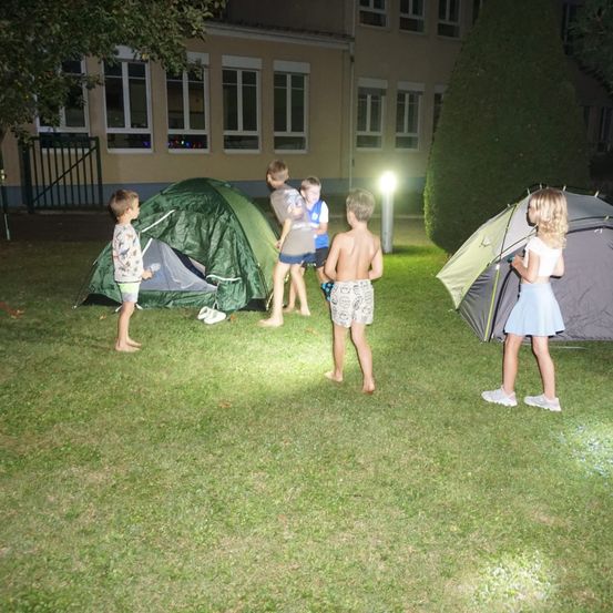 Eine Gruppe von Kindern spielt nachts im Hof, mit zwei aufgestellten Zelten und einer Lampe, die Licht spendet. Sie stehen in der Nähe der Zelte und scheinen Spaß zu haben.