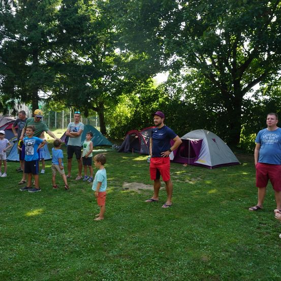 Eine Gruppe von Menschen steht auf einer Rasenfläche, einige tragen Hüte und Turnschuhe, mit Zelten im Hintergrund. Bäume und ein Zaun sind sichtbar.