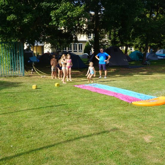 Eine Gruppe von Kindern und einem Erwachsenen spielen draußen in der Nähe von Zelten, mit einer bunten Wasserrutsche und einem Baum im Hintergrund.
