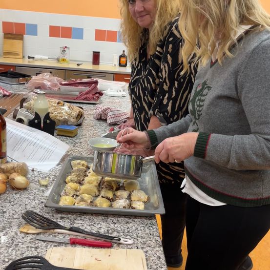 Zwei Frauen in einer Küche, eine gießt Sauce über Essen auf einem Tablett, die andere beobachtet. Verschiedene Zutaten und Utensilien sind auf dem Tresen.