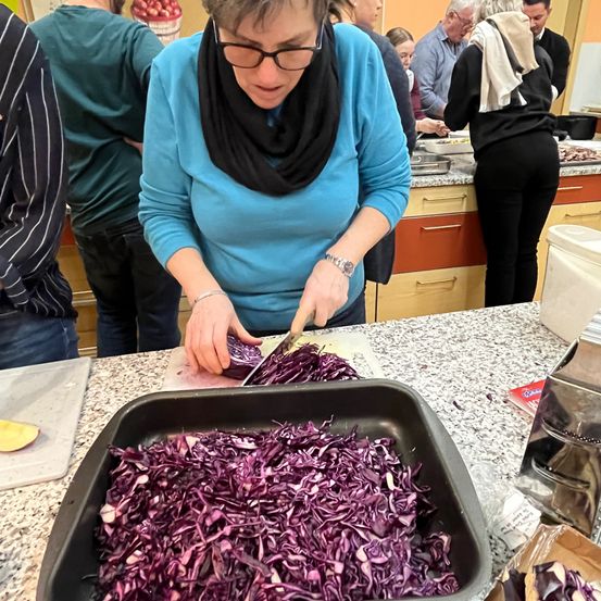 Eine Frau in einem blauen Shirt und Brille schneidet in einer Küche rotkohl. Andere Leute stehen hinter ihr, und eine Schüssel mit Essen ist auf dem Tresen.