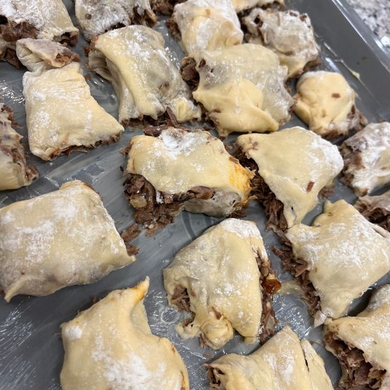 Ein Tablett mit teilweise gebackenen, blättrigen Teigtaschen, gefüllt mit einer Mischung aus Fleisch und Käse, bestreut mit Mehl, die eine rustikale und appetitliche Präsentation zeigen.