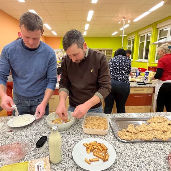 Zwei Männer bereiten in einer Küche mit anderen Essen zu. Sie haben Schüsseln, Teller und Tabletts mit Essen auf einem Tresen. Im Hintergrund befinden sich Schränke und Fenster.