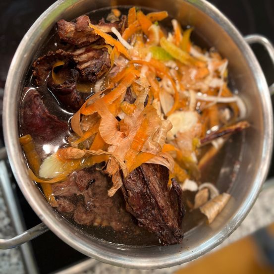 Ein Topf mit einer braunen Flüssigkeit, möglicherweise Brühe, gefüllt mit Fleischstücken, Zwiebeln und Karotten.