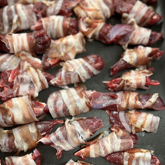 Eine Nahaufnahme von mehreren in Speck eingewickelten Fleischstücken, wahrscheinlich Hühnerlebern, auf einer schwarzen Oberfläche.