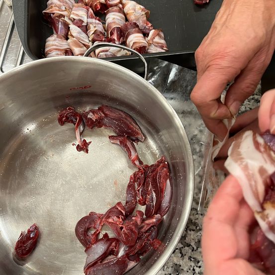 Eine Person wickelt Fleisch in Speck ein, mit rohem Fleisch in einem Edelstahltopf und Speck-umwickeltem Fleisch auf einem Tablett.