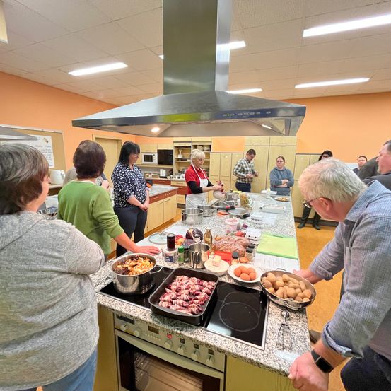 Eine Gruppe von Menschen in einer Küche kocht verschiedene Lebensmittel, darunter Eier und Kartoffeln. Eine Person rührt einen Topf auf dem Herd, während andere zusehen.
