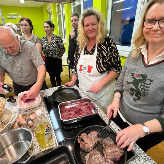 Eine Gruppe von Menschen befindet sich in einer Küche und bereitet Essen zu. Eine Frau in einer weißen Schürze hält einen Kochlöffel, und ein Mann schneidet Fleisch. Andere Menschen stehen in der Nähe und beobachten. Die Küche hat grüne Wände, einen Gasherd und verschiedene Zutaten auf der Arbeitsplatte.