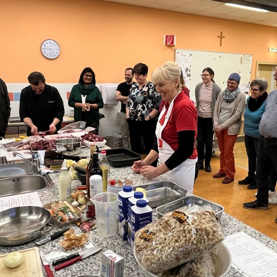Eine Gruppe von Menschen befindet sich in einer Küche mit einem Tisch voller Zutaten. Eine Frau lächelt und bereitet Essen zu. Es gibt Töpfe, Pfannen und Flaschen auf dem Tisch. Andere Menschen stehen und beobachten. Eine Uhr und ein Kreuz befinden sich an der Wand.