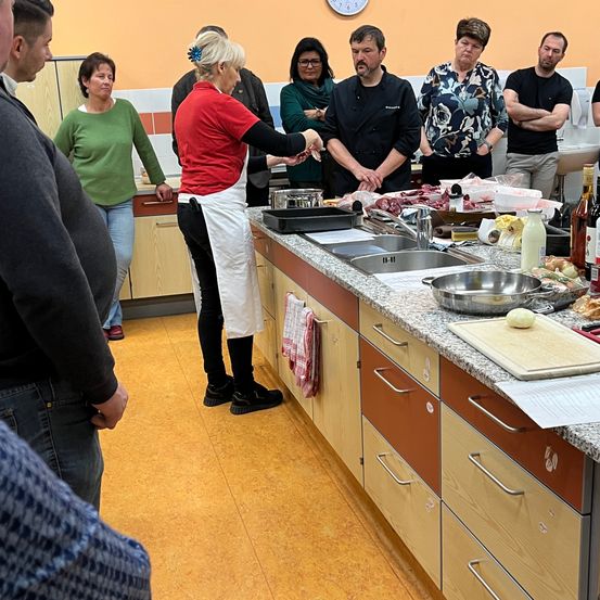 Eine Gruppe von Menschen in einer Küche, mit einer Frau in einem roten Shirt, umgeben von Zutaten, einem Waschbecken und einem Schneidebrett. Eine Uhr ist an der Wand.
