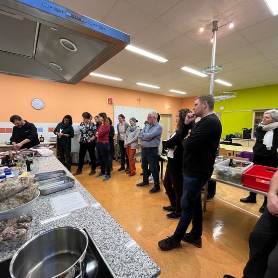 In einer Küche steht eine Gruppe von Menschen um einen Tisch mit verschiedenen Lebensmitteln und Behältern. Ein Mann spricht, und andere hören zu. Der Raum hat Holzböden und Wände mit einer Uhr und einem Kreuz.