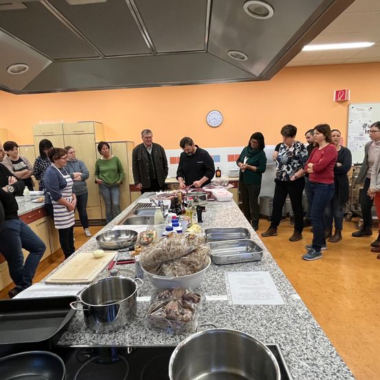 Eine Gruppe von Menschen versammelt sich in einer Küche und beobachtet, wie ein Mann auf einem langen Tisch Zutaten vorbereitet. Der Tisch ist mit Schüsseln, Flaschen und verschiedenen Zutaten bestückt. Sie stehen herum, einige mit verschränkten Armen, und die Küche hat eine Marmorplatte, Holzschränke und eine orange Wand.