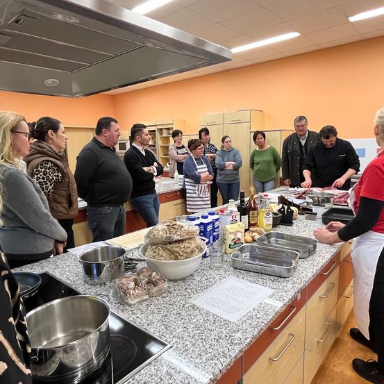 Eine Gruppe von Menschen befindet sich in einer Küche. Sie stehen um einen Arbeitstisch mit Essen und Zutaten herum. Eine Person in einem roten Shirt bereitet Essen zu. Auf dem Arbeitstisch befinden sich Kochutensilien und Zutaten. Die Küche hat einen gefliesten Boden und orangefarbene Wände.