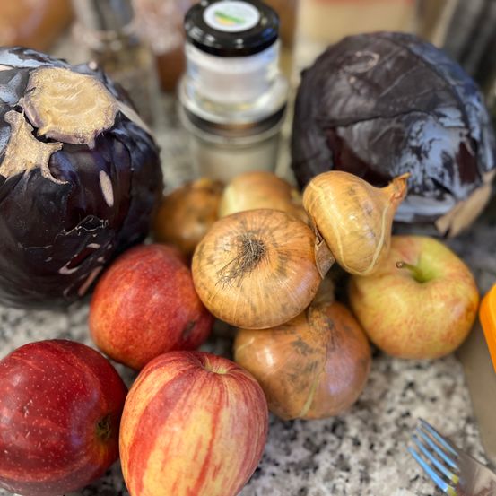 Auf einem Tisch sind verschiedene Gemüse und Früchte zu sehen, darunter Auberginen, Zwiebeln und Äpfel, zusammen mit einer Gabel und einem Gewürzstreuer.