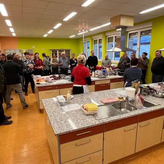 Eine Gruppe von Menschen versammelt sich in einer Küche mit Granitarbeitsplatten. Einige stehen hinter der Theke, während andere beobachten. Zutaten und Utensilien liegen auf der Theke.