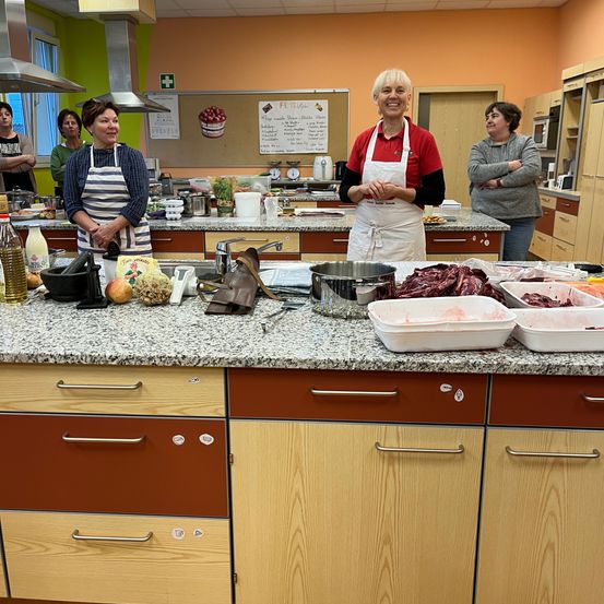 Eine Gruppe von Menschen steht in einer Küche, zwei von ihnen tragen Schürzen. Verschiedene Zutaten und Küchenutensilien sind auf dem Tresen. Eine Tafel mit Schriftzug ist an der Wand montiert.