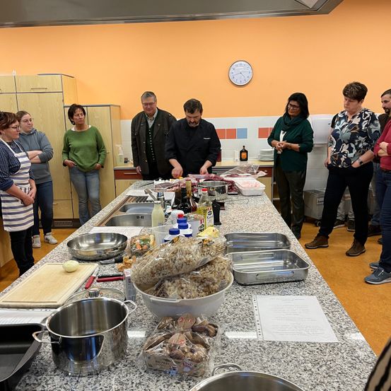 Eine Gruppe von Menschen befindet sich in einer Küche, einige beobachten und andere bereiten Essen auf der Arbeitsplatte zu. Flaschen, Schüsseln und Tabletts stehen auf dem Tisch.