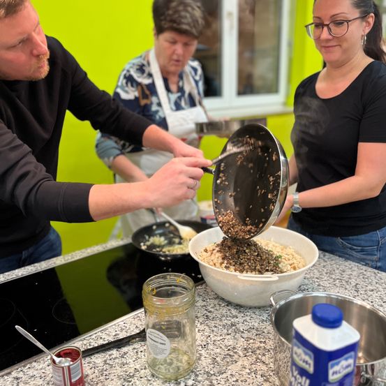 Drei Personen kochen in einer Küche. Ein Mann gießt Zutaten aus einer Pfanne in eine Schüssel. Eine Frau steht in der Nähe mit einer Schüssel. Die dritte Person steht hinter ihnen.
