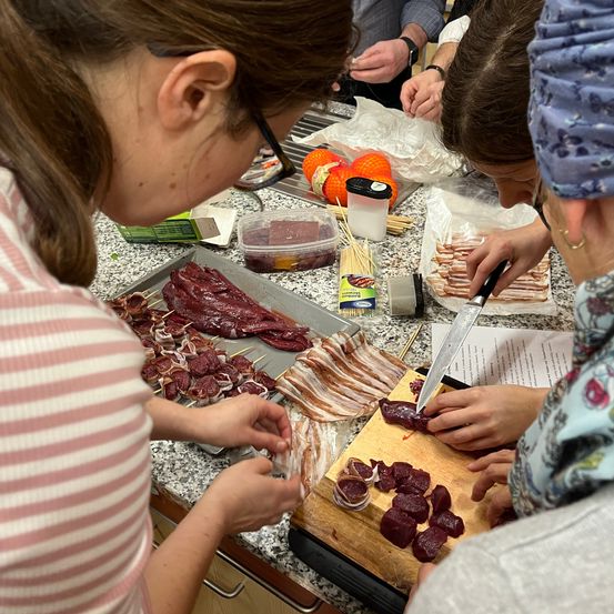 Mehrere Personen befinden sich in einer Küche und bereiten Essen zu. Eine Person schneidet Rüben auf einem Schneidebrett, während eine andere Speck schneidet. Sie haben verschiedene Zutaten und Utensilien auf der Theke.