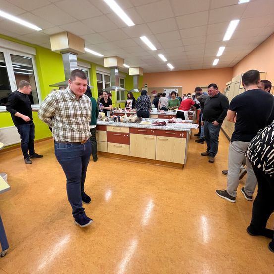 Eine Gruppe von Menschen befindet sich in einer Küche, einige stehen herum und andere arbeiten an einer Theke. Die Küche hat weiße Wände, gelbe Akzente und einen Holzboden.