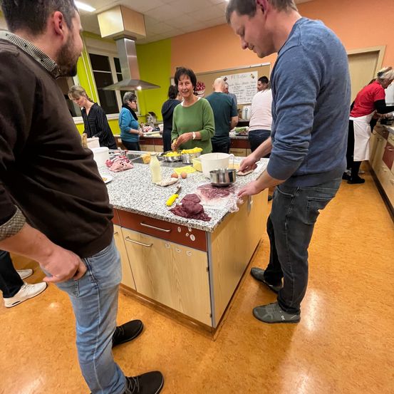Eine Gruppe von Menschen in einer Küche bereitet Essen zu. Ein Mann in einem blauen Hemd mischt Zutaten auf einer Arbeitsplatte. Eine Frau in einem grünen Hemd lächelt und beobachtet.