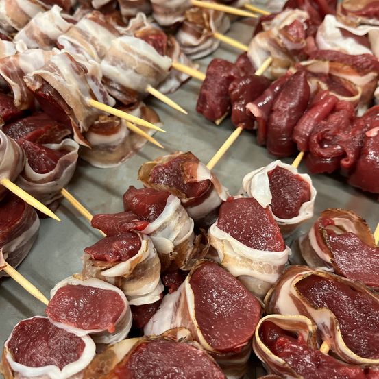 Nahaufnahme verschiedener Fleischspieße, in Speck eingewickelt, auf einem Tisch. Die Spieße sind hölzern, und das Fleisch ist rot und leicht glänzend. Der Speck scheint die Fleischstücke vollständig zu bedecken.
