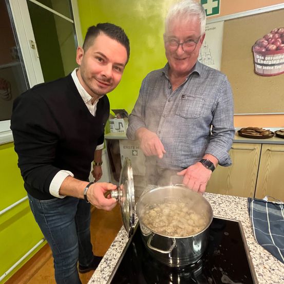 Zwei Männer kochen in einer Küche. Einer hält einen Topfdeckel, während der andere den Topf rührt. Es gibt einen Herd, ein Schneidebrett und eine Schüssel in der Nähe.