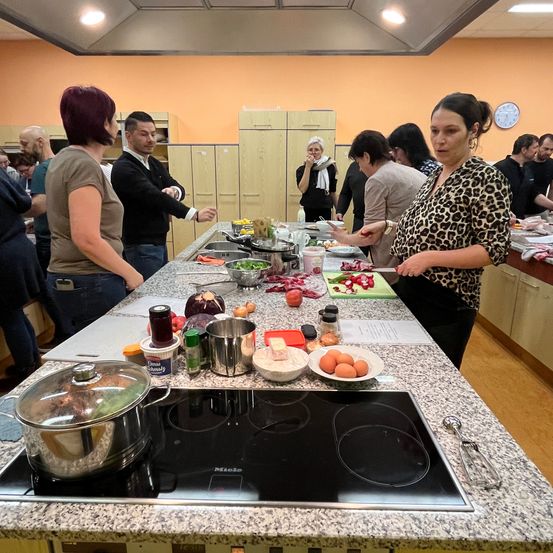 Eine Gruppe von Menschen ist in einer Küche und bereitet Essen zu. Zutaten und Utensilien sind auf dem Tresen. Eine Frau steht und spricht mit anderen.