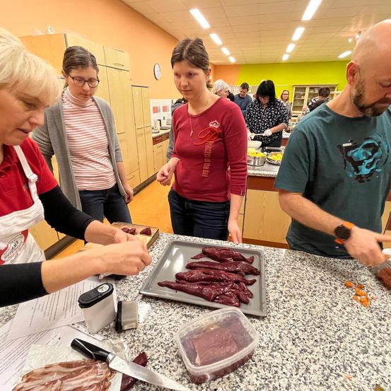Eine Gruppe von Menschen ist in einer Küche und bereitet Essen zu. Eine Frau in einem roten Shirt zeigt auf eine Fleischschale. Eine weitere Frau in einer grauen Jacke hält ein Messer. Ein Mann in einem grünen Shirt hält eine Schüssel. An der Wand hängt eine Uhr.