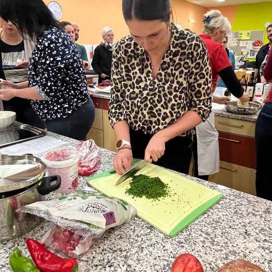 Eine Frau in einem Leopardenmusterhemd schneidet grüne Kräuter auf einem Schneidebrett, während andere im Hintergrund Essen zubereiten.