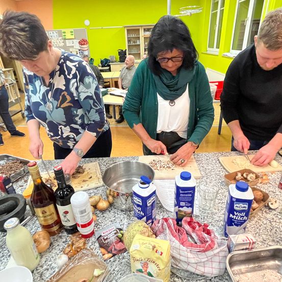 Drei Personen arbeiten in einer Küche und schneiden Gemüse. Der Tisch ist mit verschiedenen Zutaten wie Flaschen, Schüsseln und Behältern gedeckt. Im Hintergrund sind weitere Personen zu sehen, und der Raum hat eine grüne Wand und einen Holzboden.