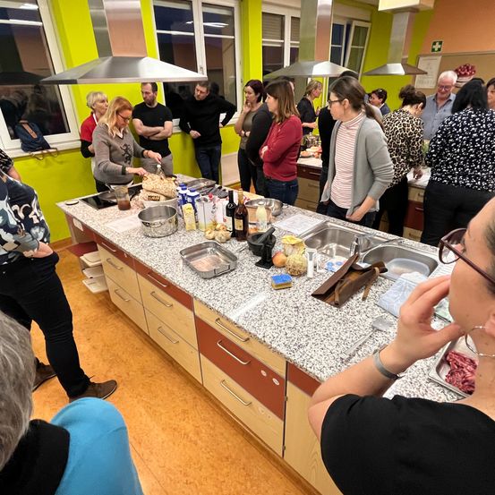 Eine Gruppe von Menschen befindet sich in einer Küche. Einige beobachten eine Person, die am Tresen kocht. Verschiedene Gegenstände stehen auf dem Tresen, darunter Flaschen, eine Pfanne und eine Schüssel.