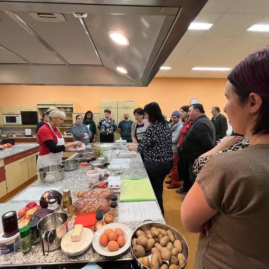 Eine Gruppe von Menschen versammelt sich um einen Küchentresen mit verschiedenen Lebensmitteln. Eine Frau in einer roten Schürze steht am Tresen und scheint Essen zuzubereiten.