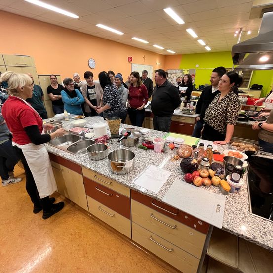 Eine Gruppe von Menschen befindet sich in einer Küche und bereitet Essen zu. Eine Frau in einer weißen Schürze steht an einem Tresen mit verschiedenen Zutaten. Andere stehen um sie herum und beobachten.