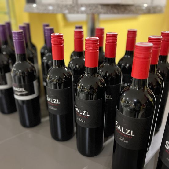 Mehrere Weinflaschen mit roten Verschlüssen stehen in einer Reihe auf einer weißen Oberfläche, mit einem gelben Hintergrund. Die Weinflaschen haben schwarze Etiketten.