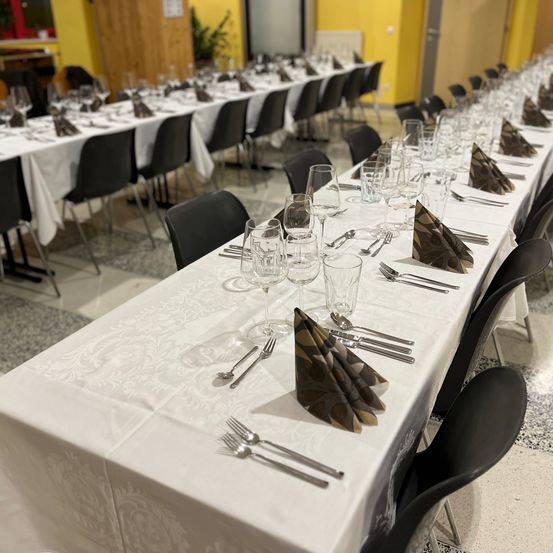 Ein Speisesaal mit mehreren langen Tischen, die mit Weingläsern, Besteck und Servietten gedeckt sind. Jeder Tisch hat Stühle auf beiden Seiten. Der Raum hat gelbe Wände und einen gefliesten Boden.